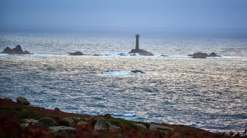 This landscape photograph captures the misty coast of Cornwall in the early evening during autumn. The main subject is Longships mist, with the Longships lighthouse prominently emerging from a cluster of rocky outcrops in the sea. The coastline, featuring rugged terrain and patches of vegetation, can be seen in the foreground, while the waves of the Atlantic Ocean reflect the dimming light. The Longships lighthouse serves as a vital landmark for maritime navigation along the Cornish coast, and the muted tones of the water and sky highlight the atmospheric conditions characteristic of autumn evenings in Cornwall.